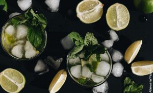 Foto de dos vasos sobre una mesa con trozos de limón, hielo y hierbabuena que muestra los mojitos y su receta