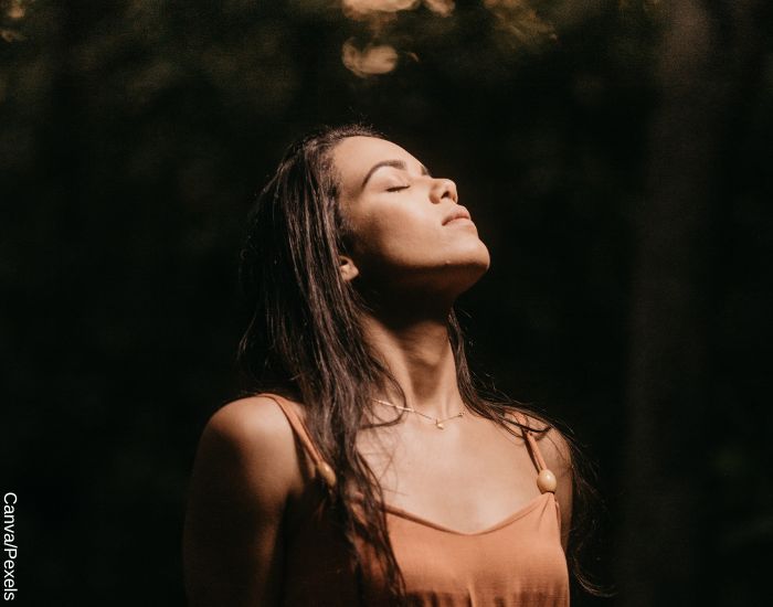 Foto de una mujer tranquila, con los ojos cerrados en medio de bosque recibiendo el sol en la cara
