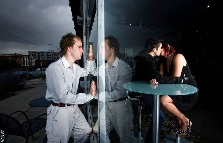 Foto de un hombre viendo a través de una ventana a una pareja besándose