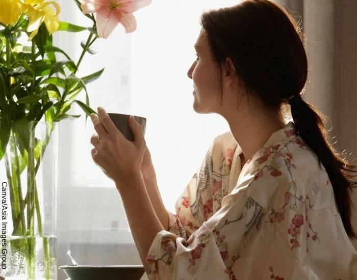Foto de una mujer en pijama tomando té sentada mientras mira por la ventana