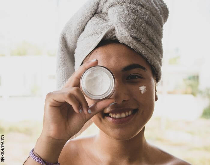 Foto de una mujer sonriente con una toalla en su cabello, sosteniendo una lata de pomada frente a su rostro
