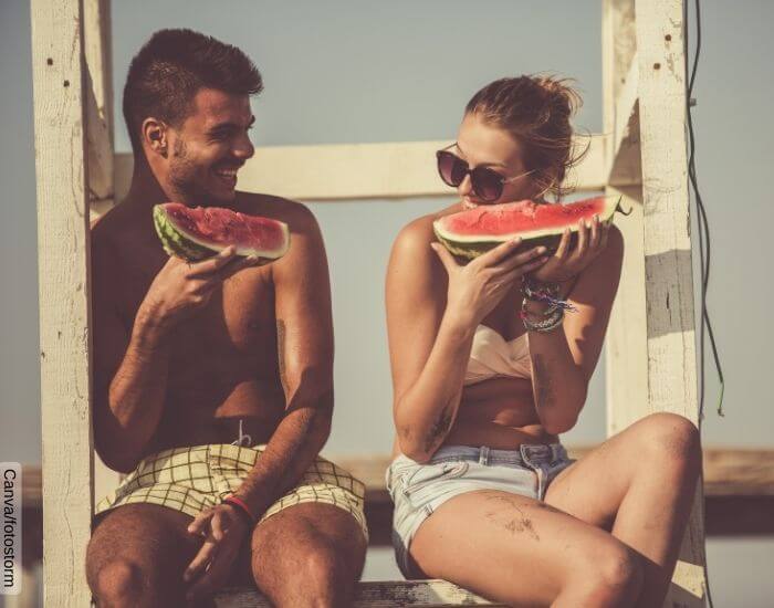 Foto de una pareja comiendo sandía en la playa sentados sobre unas escaleras