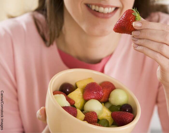 Foto de una mujer sonriendo mientras come un bowl de fruta con fresas, piña y uvas