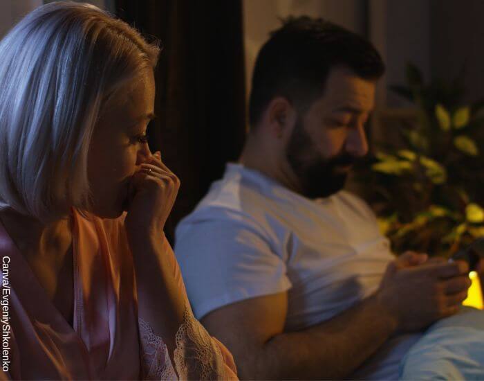 Foto de una pareja en la cama antes de dormir, el hombre mira el celular mientras la mujer está preocupada