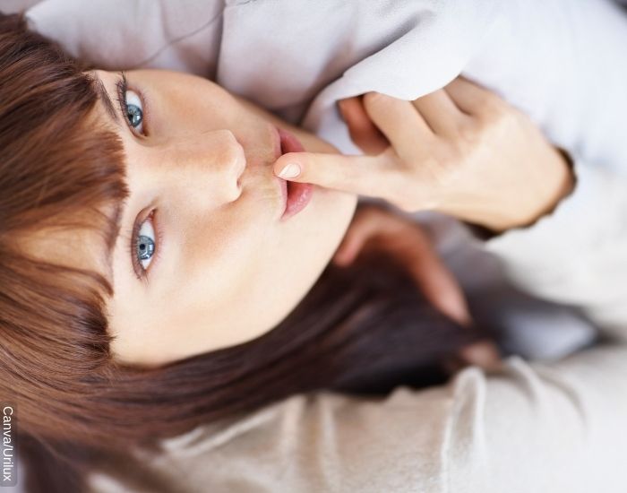 Foto de una mujer recostada sobre la cama con su índice frente a los labios indicando que guarda un secreto