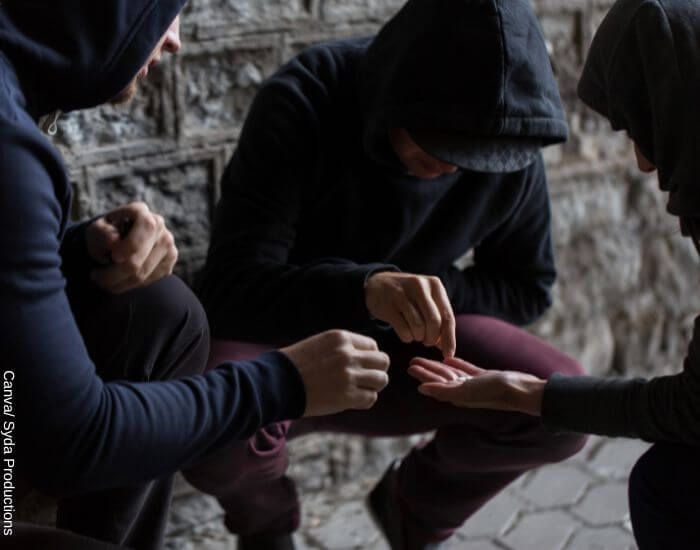 Foto de tres hombres con capota agachados en una esquina compartiendo pastillas entre ellos