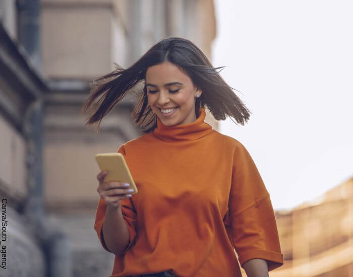 Foto de una mujer caminando por la calle mientras mira el celular sonriendo por tener química con una persona que le está escribiendo