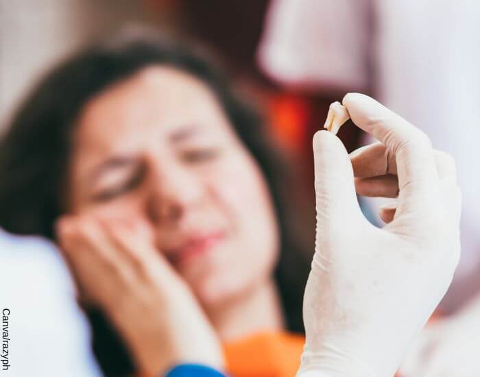 Foto de una mano con un guante de latex sosteniendo una muela adulta, una mujer al fondo se toca la mejilla en señal de dolor