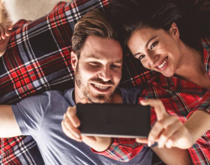 Foto de una pareja posando para tomarse fotos con el celular recostados en la cama