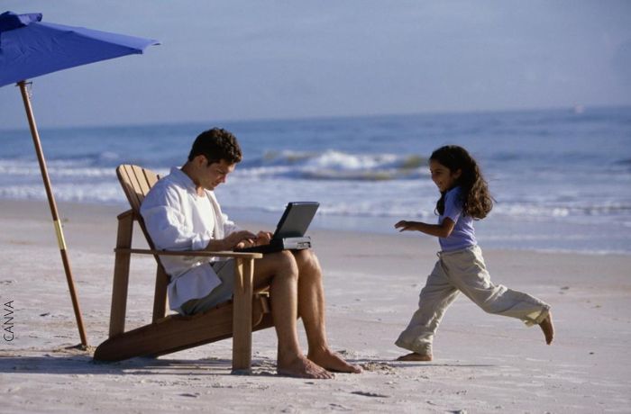 Foto de un hombre en la playa escribiendo en el computador y con una niña cerca