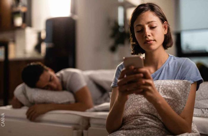 Foto de una mujer escribiendo en el celular mientras su pareja está dormida detrás