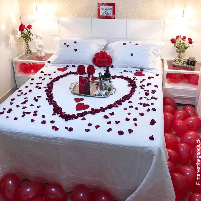 Foto de una cama con un corazón con petalos de rosas y un vino en la mitad