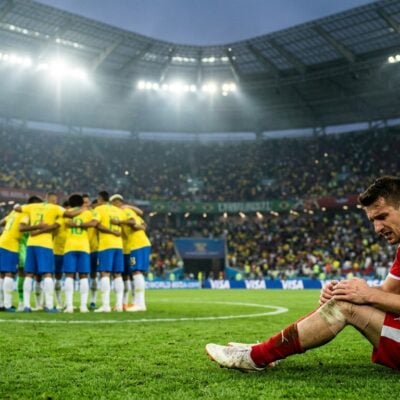 Un futbolista de rojo se lamenta sentado en el césped mientras el equipo rival de amarillo celebra al fondo