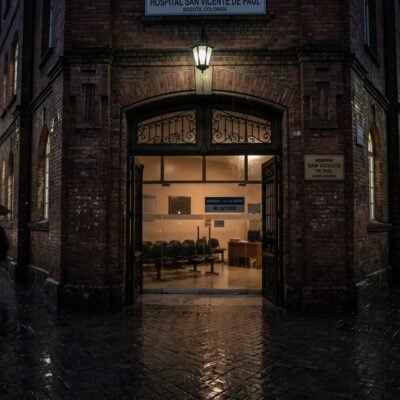 Fachada del Hospital San Vicente de Paúl en Bogotá durante una noche lluviosa, con peatones bajo paraguas